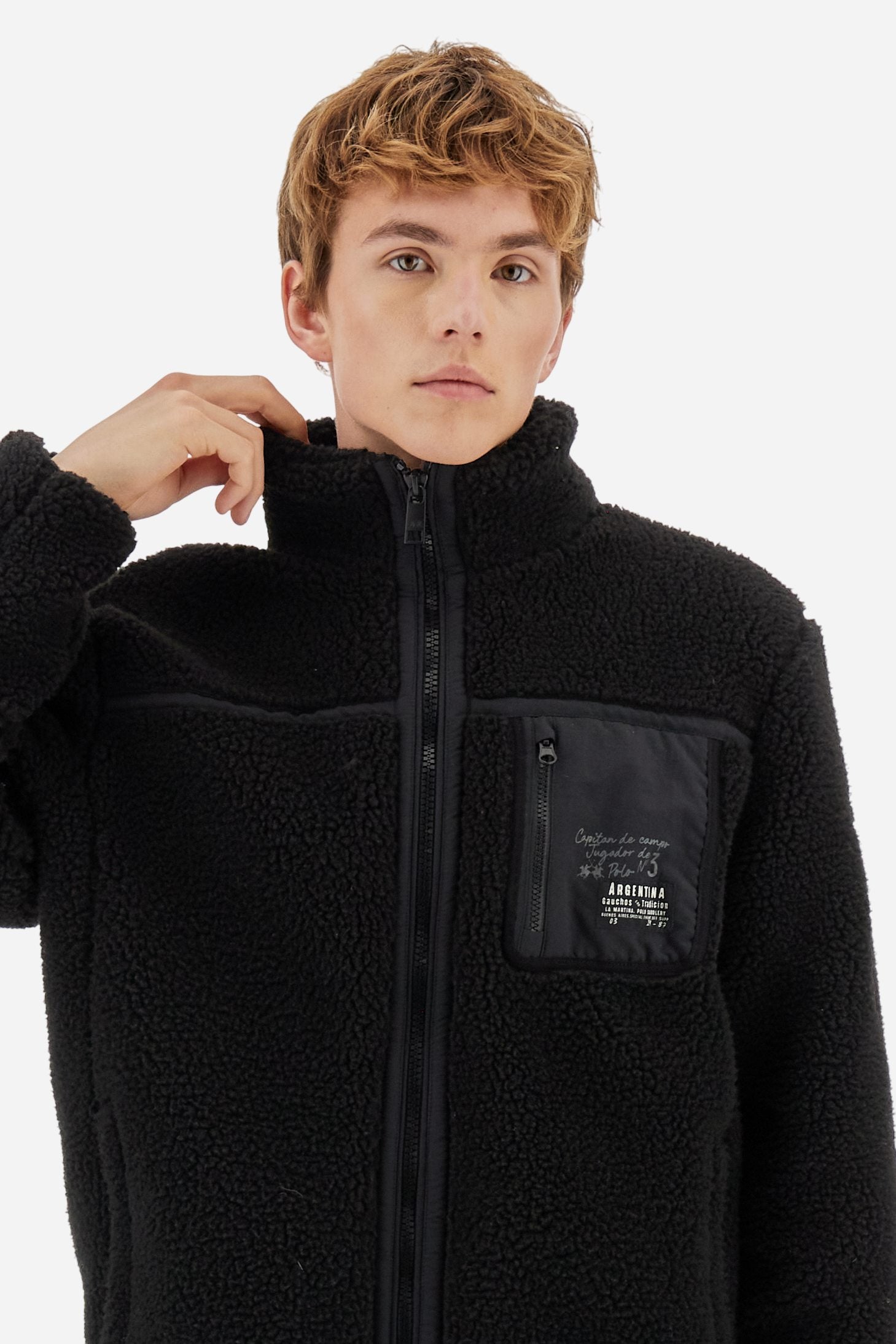 Men’s teddy jacket in 100% polyester with high collar and full lining. Front closure with customized tone-on-tone zip. Finished with matte nylon trims on seams and edges, also used for the zipped chest pocket. High collar offers extra wind protection. Patches inspired by Argentina and Jugador de Polo on sleeve and chest pocket. Regular, straight fit. Model wears size L. - Black
