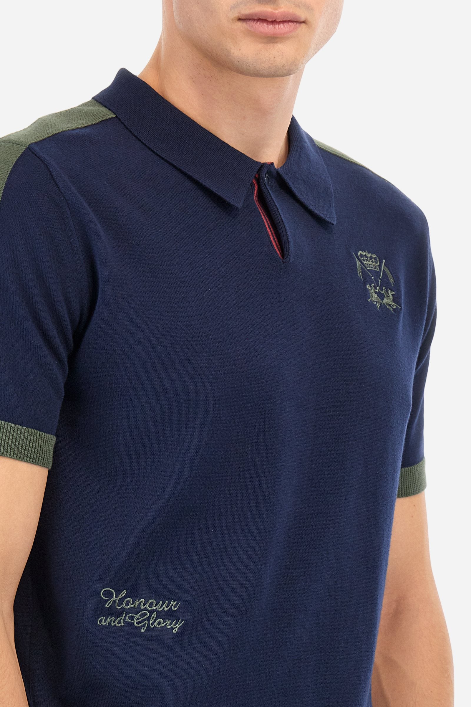 Men's two-tone short-sleeved knitted polo shirt made of 100% cotton. The cuffs are contrasting, while the waistband is in the same tone, but both are ribbed. The applied bands on the sleeve and shoulders are knitted in a contrasting colour. Contrasting details are present on the neck opening and inside the collar as a decorative motif. Small embroidered lettering on the front. The logo is embroidered in a contrasting colour on the chest, left side. The fit is regular. The model is wearing a size L. - Navy
