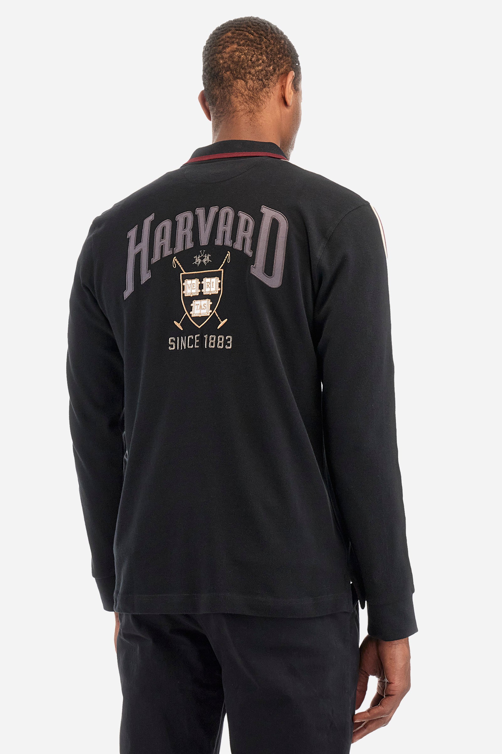 Men's plain short-sleeved polo shirt, made from 100% cotton. Half-zip fastening. Trimmed collar. The sleeves are finished with contrasting stripes. The symbols and lettering associated with the Harvard line are appliquéd on the back. The front is embroidered with the logo and La Martina lettering. Regular fit. The model is wearing a size L. - Black