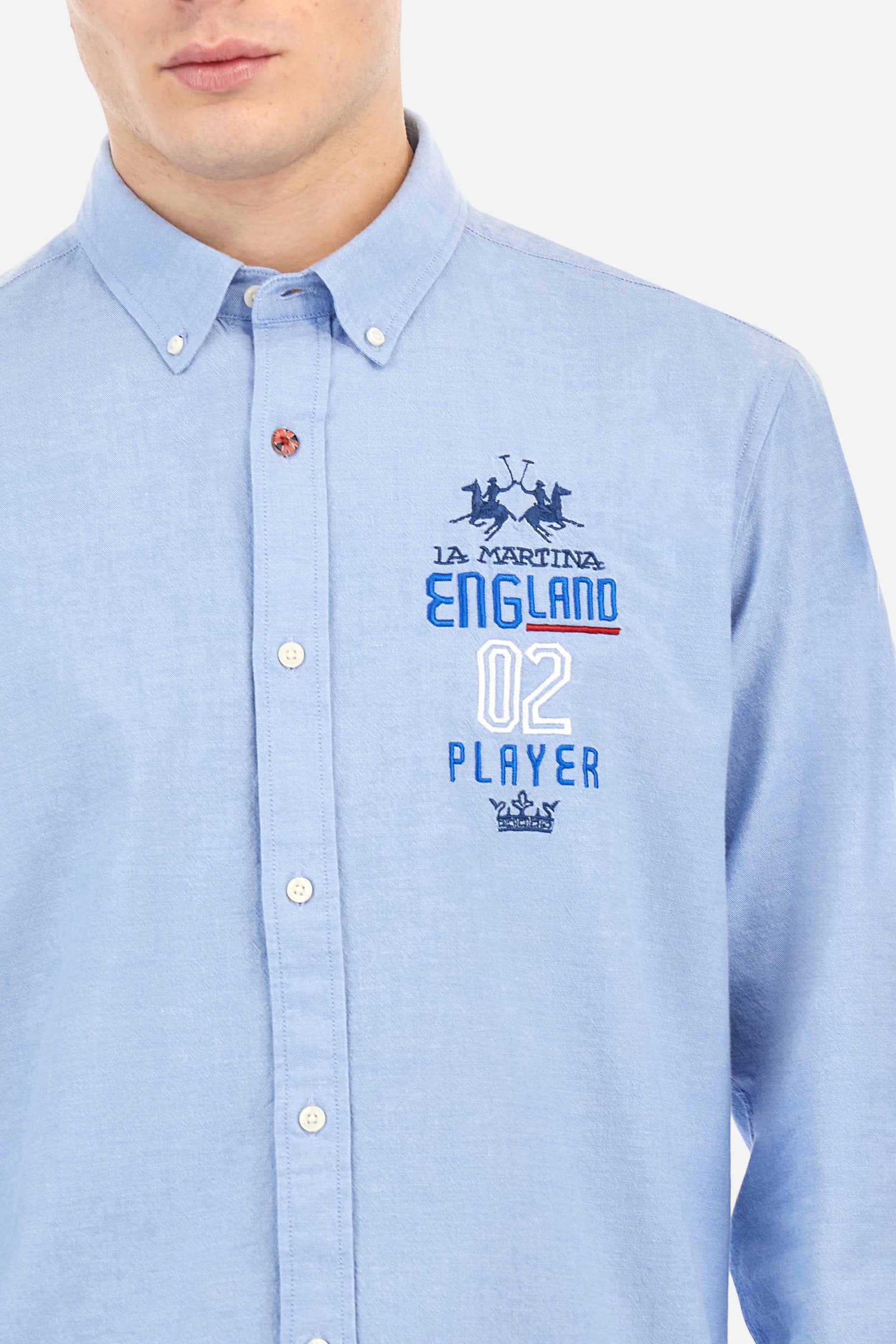 Men's long-sleeved plain shirt made from 100% Oxford cotton. The button-down collar is finished with a second button printed with the English flag. The multicoloured embroidery on the chest, over the heart, includes lettering, numbers and the logo, which add a distinctive touch. Regular fit. The model is wearing a size L. - Cornflower blue