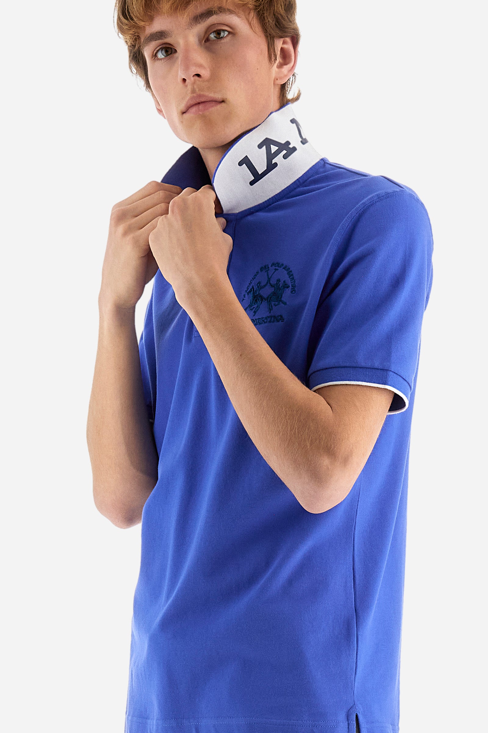 Men's plain short-sleeved polo shirt made from stretch piqué. Featuring a two-button fastening and a ribbon on the inside collar in contrasting coloured grosgrain. The collar and cuffs are in matching rib knit, with the undercollar also in rib knit but in a contrasting colour. The large logo is embroidered on the chest, left side with contrasting coloured thread. The undercollar features a print of the La Martina lettering in a contrasting colour. Regular fit. The model is wearing a size L. - Dazzling Blue