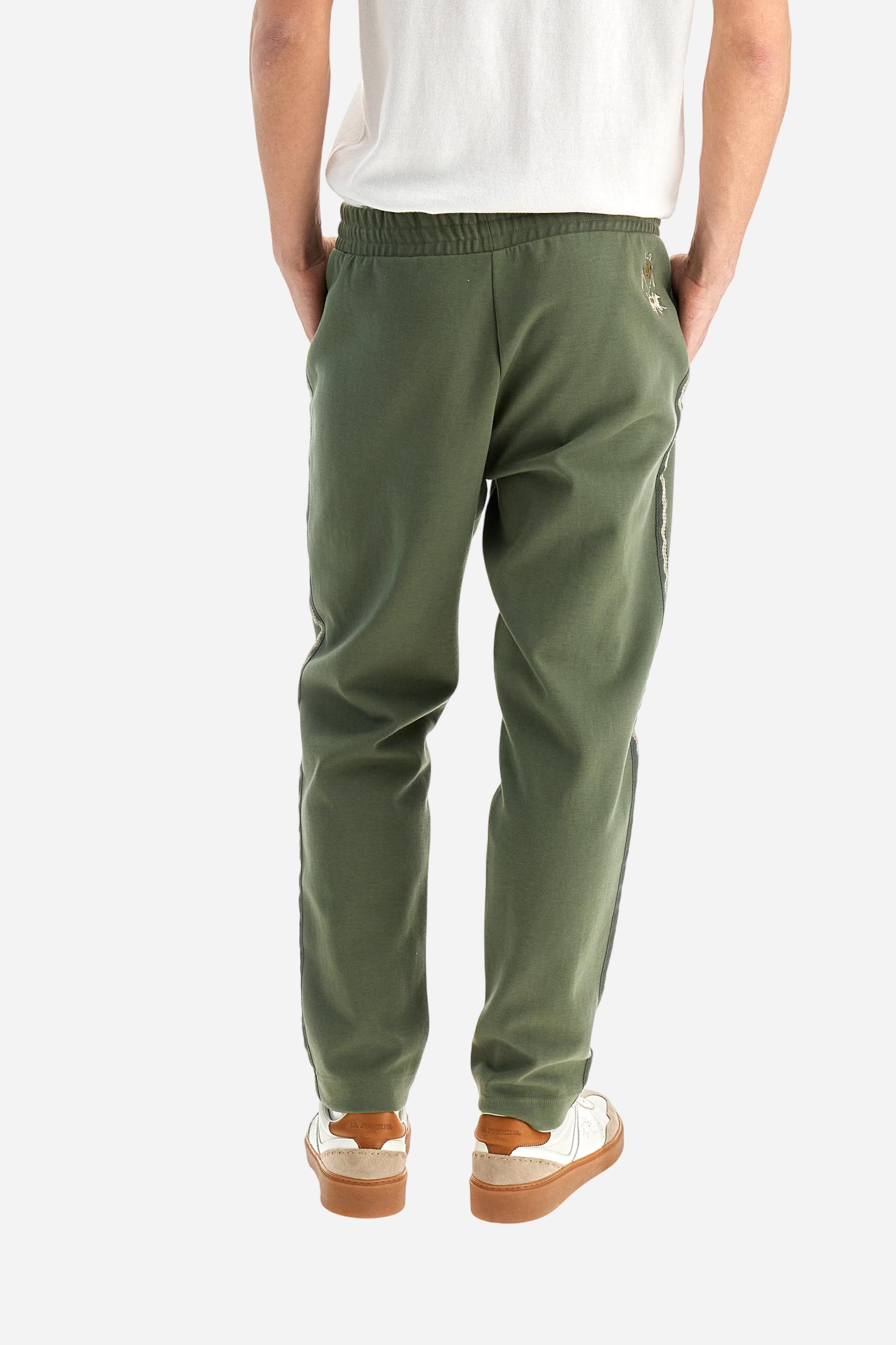 Single-colour men's jogging trousers made of lightweight cotton-blend interlock fleece. Drawstring waistband. Side pockets with matching zip fastening. On both sides, there are multicoloured striped jersey bands. The front features vertical ribbed detailing. The logo is embroidered in colour-contrasting thread on the back, under the strap, towards the right side. Regular fit. The model is wearing a size L. - Thyme
