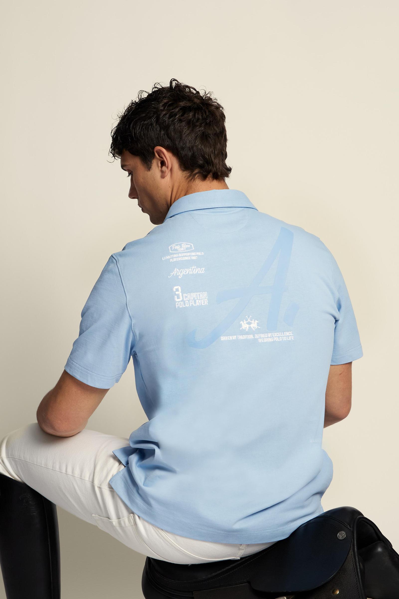 Men’s heavy cotton jersey polo with three-button placket and striped details. Celebratory maxi graphic and bold lettering. - Blue Bell
