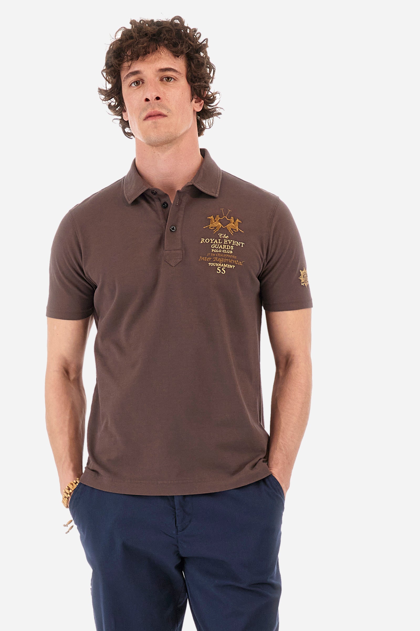 Men's short-sleeved single-colour polo shirt in elasticated cotton. Two-button fastening. Pointed collar. On the front we find copper-coloured lettering and a large logo. On one of the sleeves there is a symbol representing the Guards line. The fit is regular. The model wears a size L. - French Roast