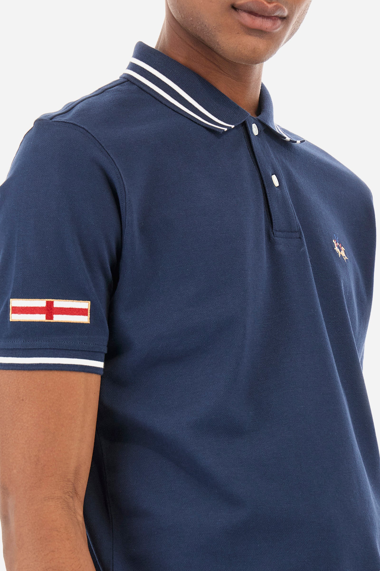 Men’s polo shirt in solid color with short sleeves in 100% cotton piquet. Two button closure. Edge and inner neck, along with the armhole all profiled with contrasting stripes. On one of the sleeves is applied a patch depicting the flag of the country to recall the capsule collection Ciudades. Small La Martina logo embroidered on the chest. The fit is regular. The model wears a size L. - Navy