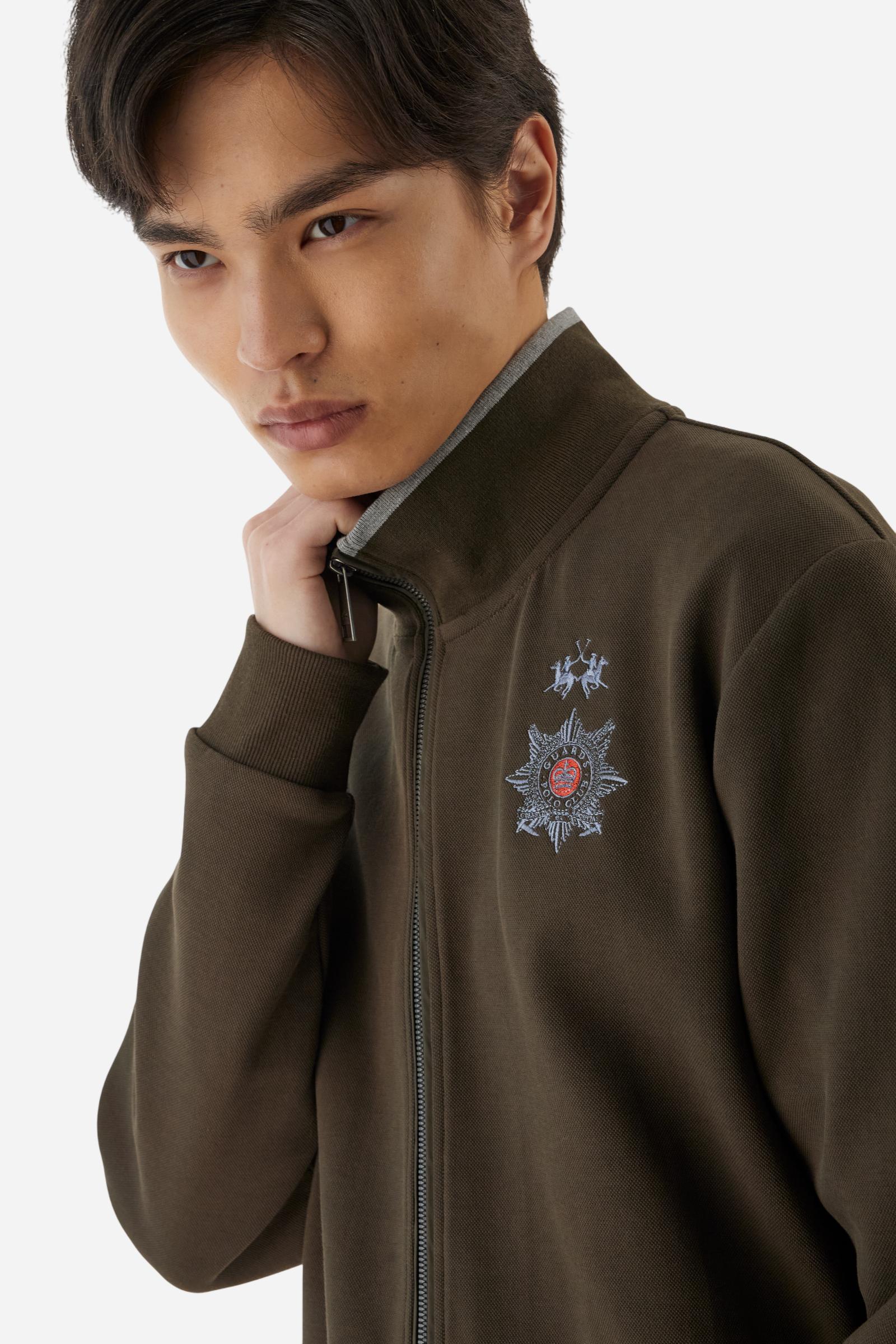 Men's long-sleeved, full-zip solid colour sweatshirt. Made of stretch cotton-blend piqué. Design with front opening fastened by a zip, high collar in two-tone ribbing. Solid colour ribbed cuffs and waistband. The small brand logo and Guards crest are embroidered in multi-coloured thread on the chest. On the right sleeve, there is another embroidery of a crest inspired by the British royal household. The regular fit silhouette is practical for everyday wear. The model is wearing a size L. - Java