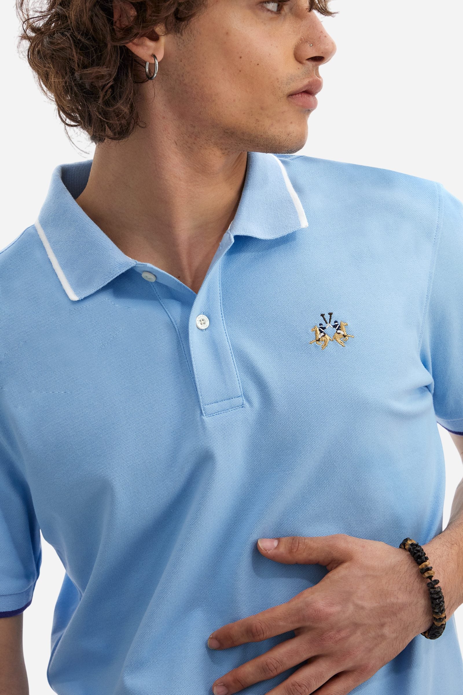 Short-sleeved, single-colour men's polo shirt made of stretch cotton piqué. Fastens with two buttons. The collar and cuffs are ribbed, and there is a colour-contrasting trim at the bottom of the collar. Inside the collar there is a grosgrain ribbon of a different shade. The logo is embroidered on the chest in a contrasting colour. The fit is slim. The model is wearing a size L. - Vista Blue