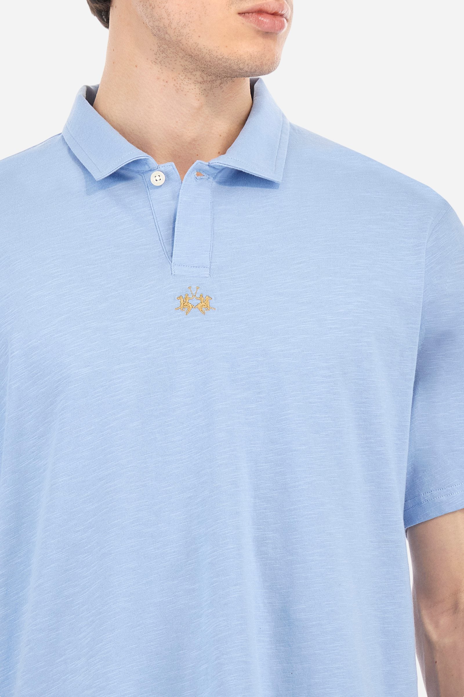 Men's short-sleeved polo shirt in a single colour, made of 100% cotton slub jersey. Fastens with a button. A fabric patch representing the garment's certificate is sewn onto the back. The logo is embroidered in contrast under the opening on the front. Comfort fit. The model is wearing a size L. - Vista Blue