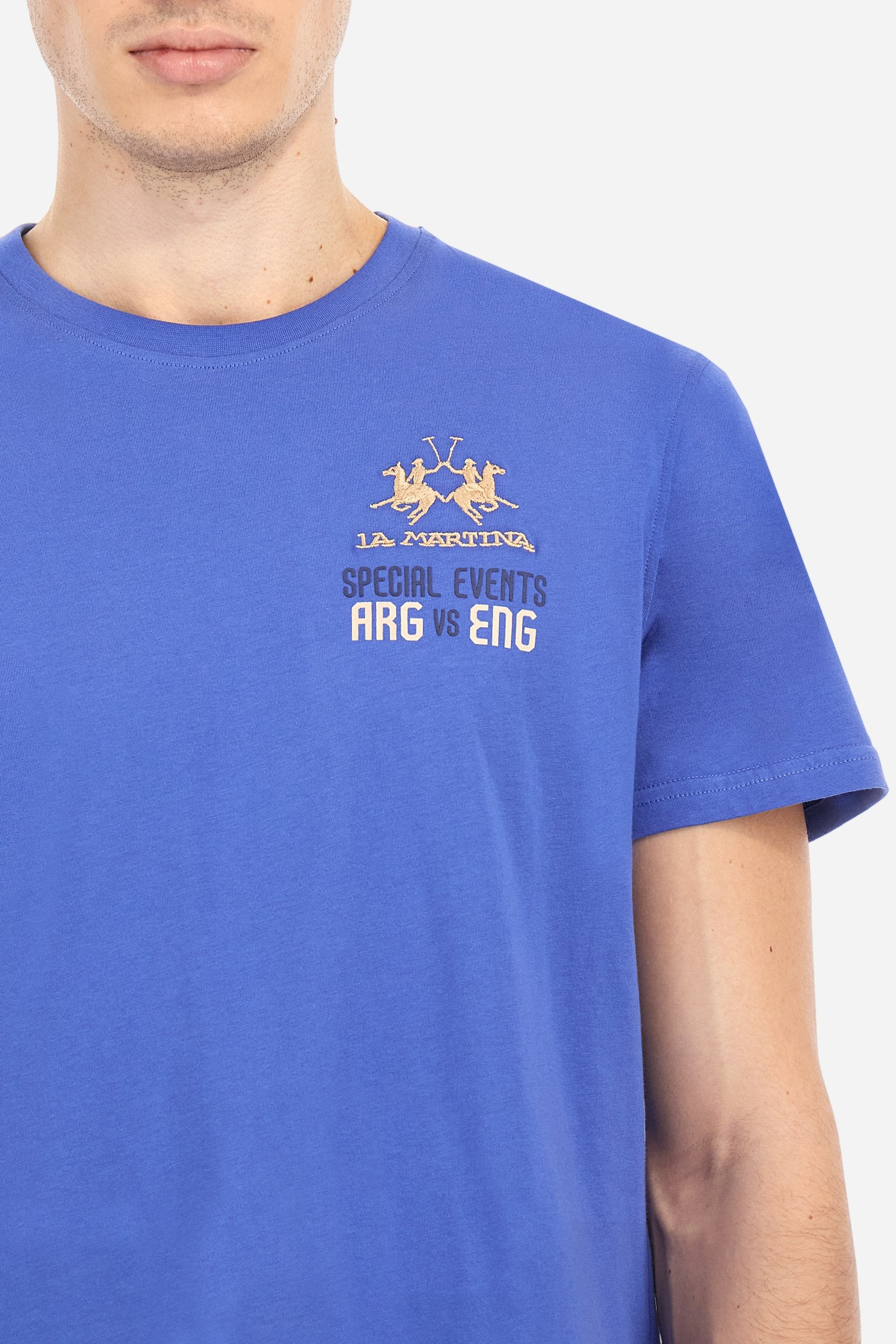 Men's plain cotton jersey short-sleeved T-shirt. Ribbed crew neck. On the front, left side, there is a two-tone embroidery inspired by the polo events played between Argentina and England. On the back, the printed graphic plays with the two flags, celebrating the Dubai and La Martina polo teams with contrasting coloured writing. The logo is embroidered on the left side of the chest in contrasting thread. Regular fit. The model is wearing a size L. - Dazzling Blue