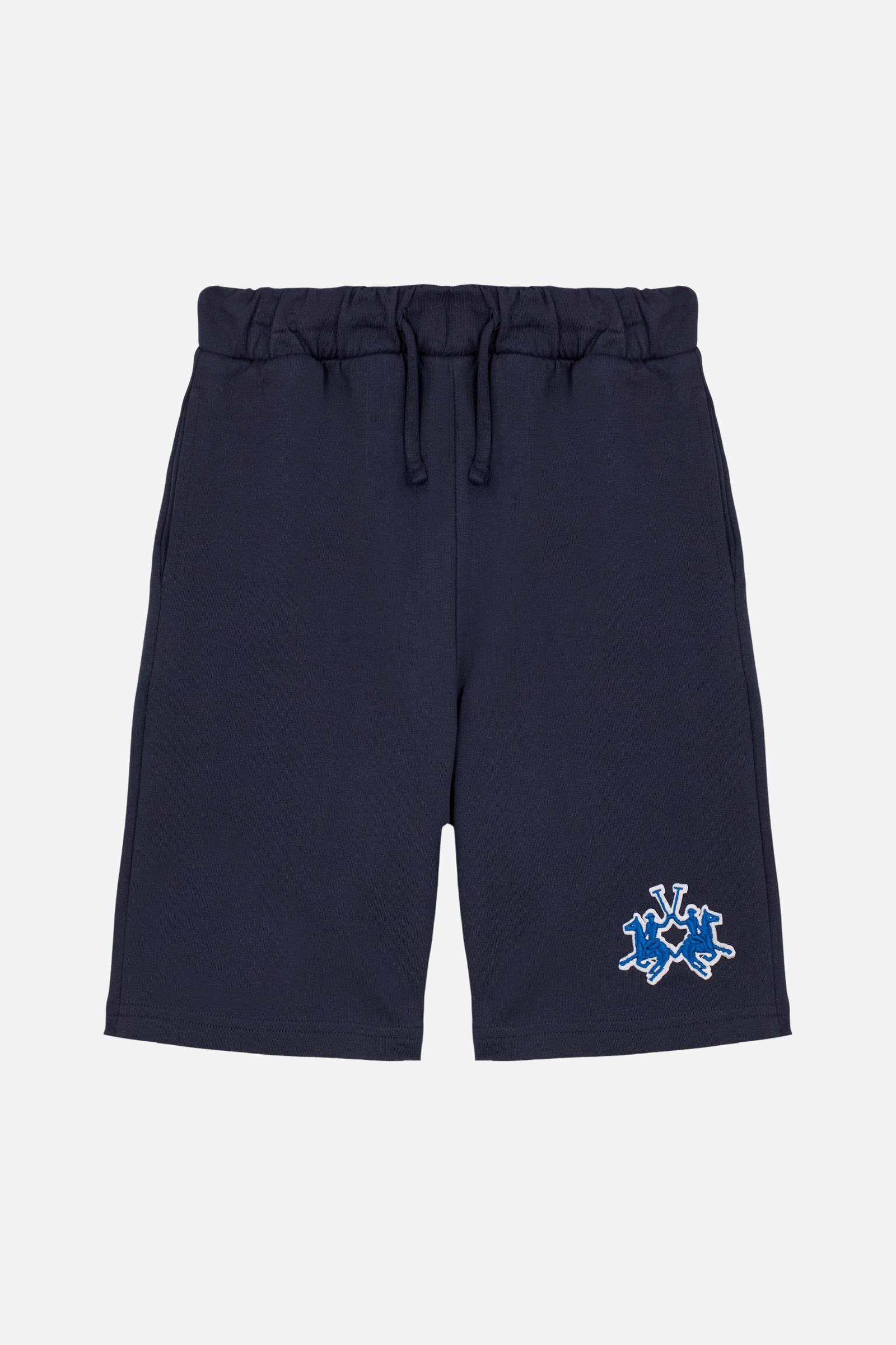Bermuda shorts for boys made from soft 100% cotton fleece in a solid colour. The style features a clean, essential design, with an elasticated waist adjustable by drawstring and practical side pockets. A patch pocket is placed on the back, while the contrast embroidered maxi logo at the leg hem adds a distinctive and recognisable detail. A comfortable, versatile garment, ideal for leisure time. - Navy
