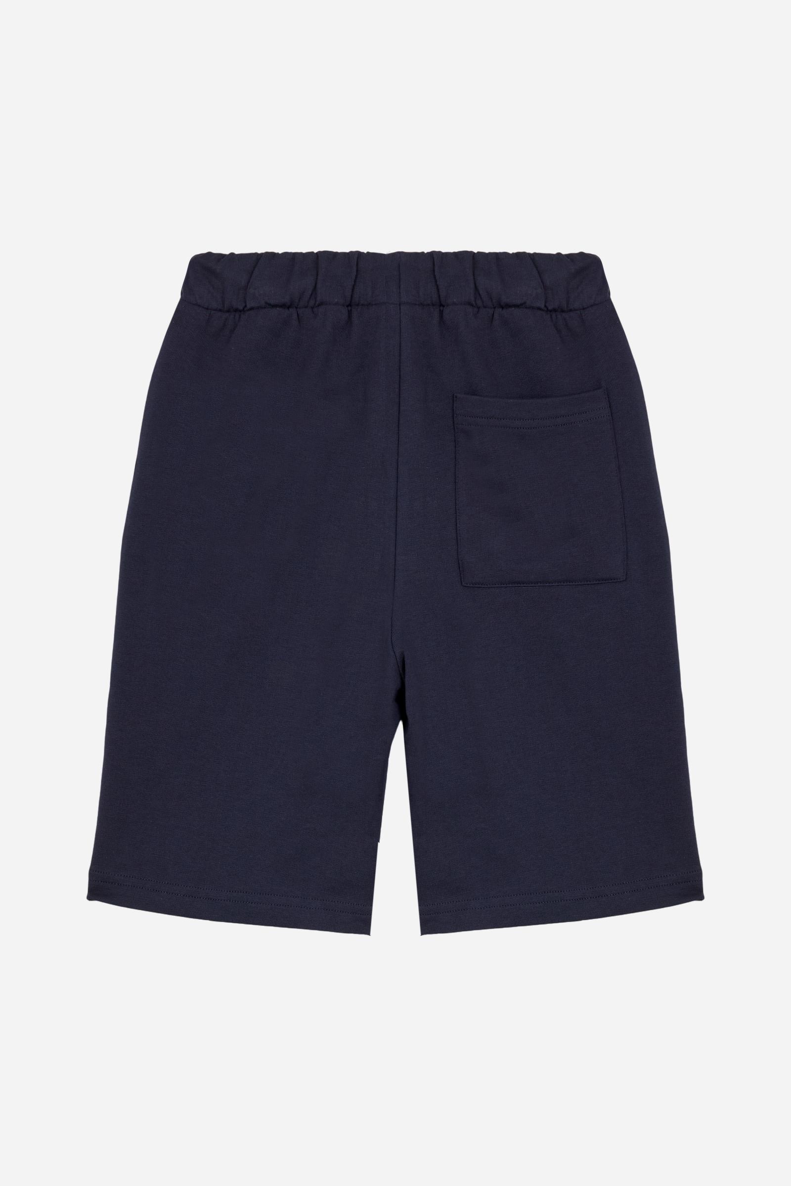 Bermuda shorts for boys made from soft 100% cotton fleece in a solid colour. The style features a clean, essential design, with an elasticated waist adjustable by drawstring and practical side pockets. A patch pocket is placed on the back, while the contrast embroidered maxi logo at the leg hem adds a distinctive and recognisable detail. A comfortable, versatile garment, ideal for leisure time. - Navy