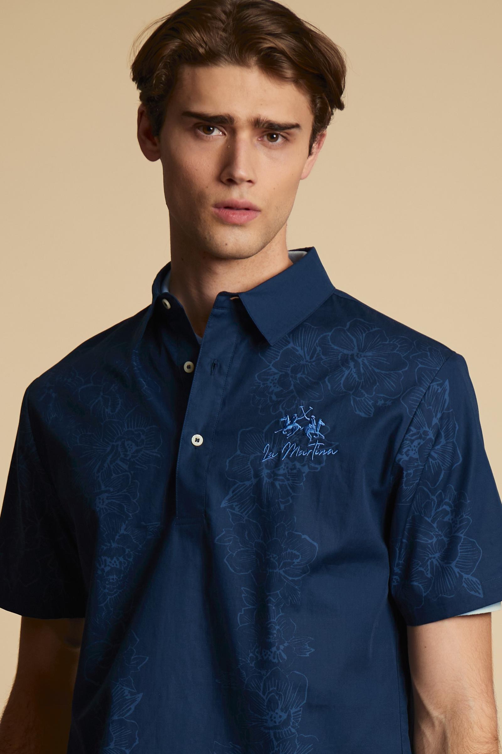 Men’s short-sleeved polo shirt made from lightweight solid-color pure cotton poplin. The contrasting floral print, with its fine graphic line, develops vertically across the front and back of the garment, adding movement to the design. The style features a classic collar and polo-style fastening with a two-button placket. The fit is comfortable, with a straight hem and short sleeves cut in a clean, roomy shape. The contrasting embroidered logo on the chest signs the garment with discretion. The model is wea