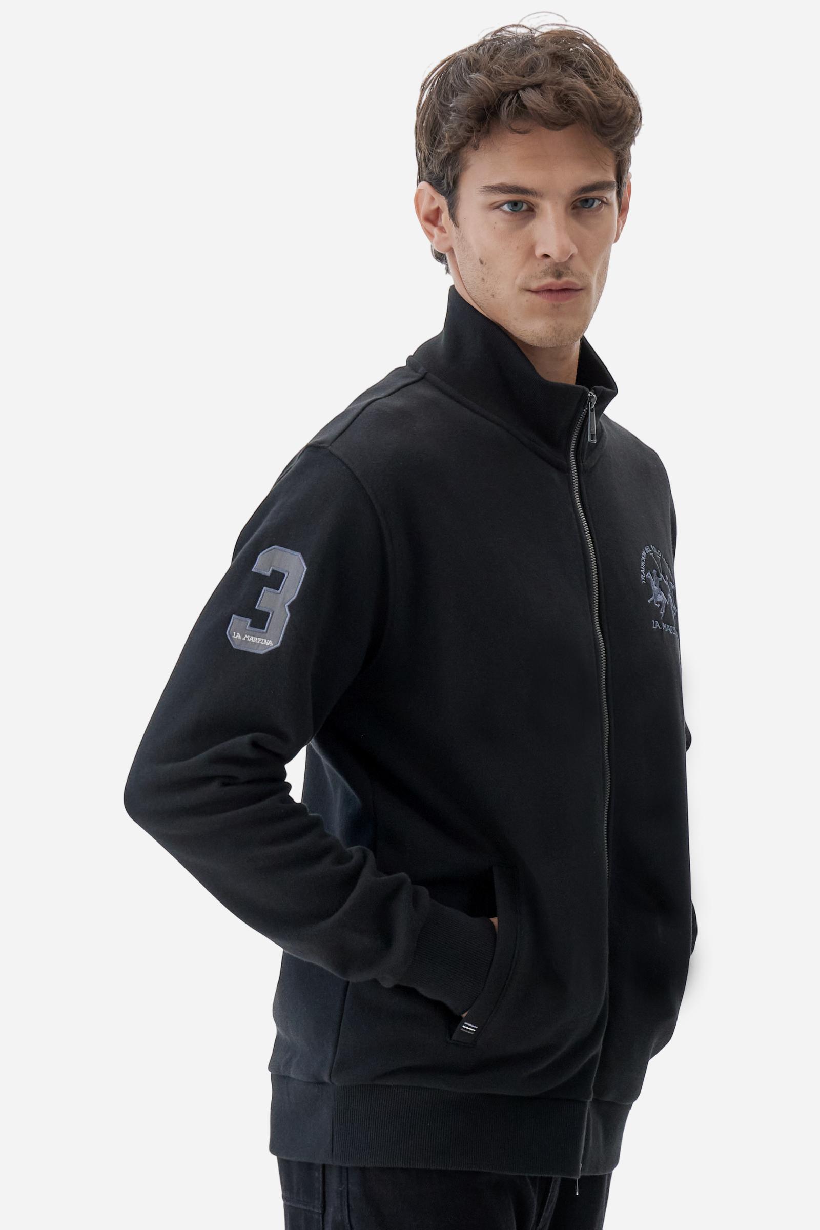 Men's long-sleeved solid colour sweatshirt. It is made of comfortable 100% cotton fleece with a soft-brushed feel. Model with high ribbed neck and front zip opening. Solid colour ribbed cuffs and waistband. The large contrasting brand team logo is embroidered on the chest. A contrasting colour fabric number patch is applied to the right sleeve. The model has a regular fit that offers a comfortable, relaxed silhouette. The model is wearing a size L. - Black